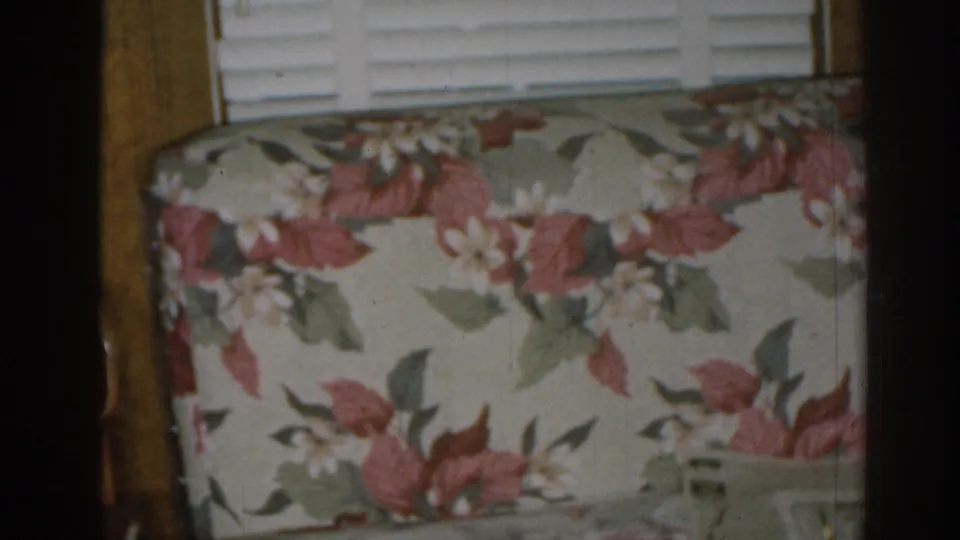 1963 New Jersey home-movie frame showing floral sofa upholstery near a window with white blinds and wood trim.