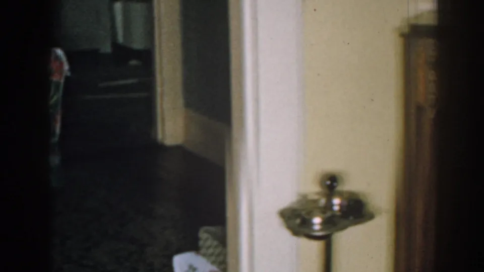 Home-movie frame from a 1963 New Jersey living room showing a doorway, dark carpet, pale wall, and small pedestal stand or ashtray.