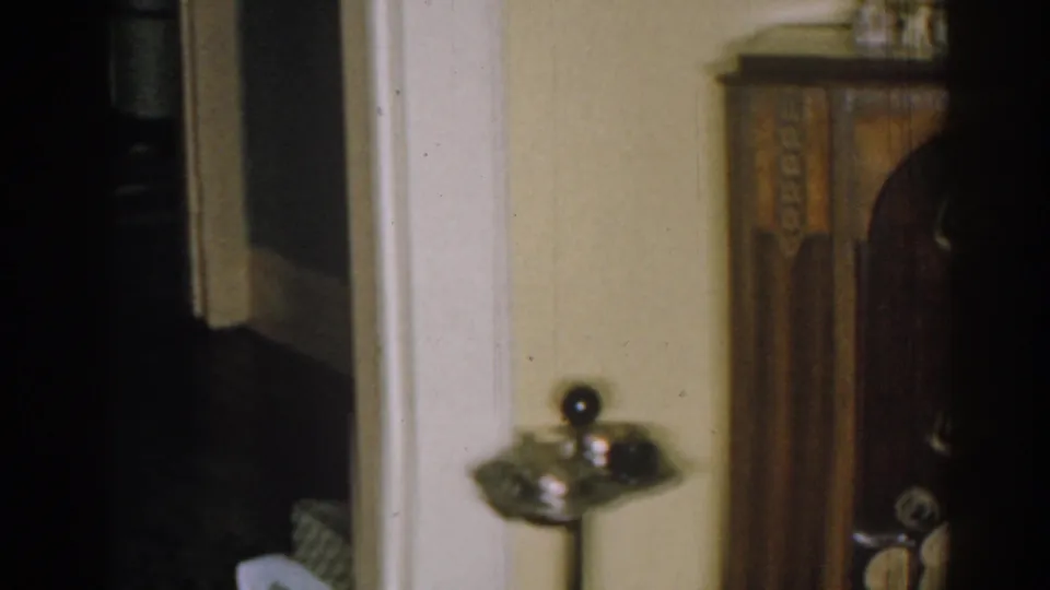 1963 New Jersey home interior with a dark carved cabinet, metal ashtray stand, pale wall, and shadowed doorway.