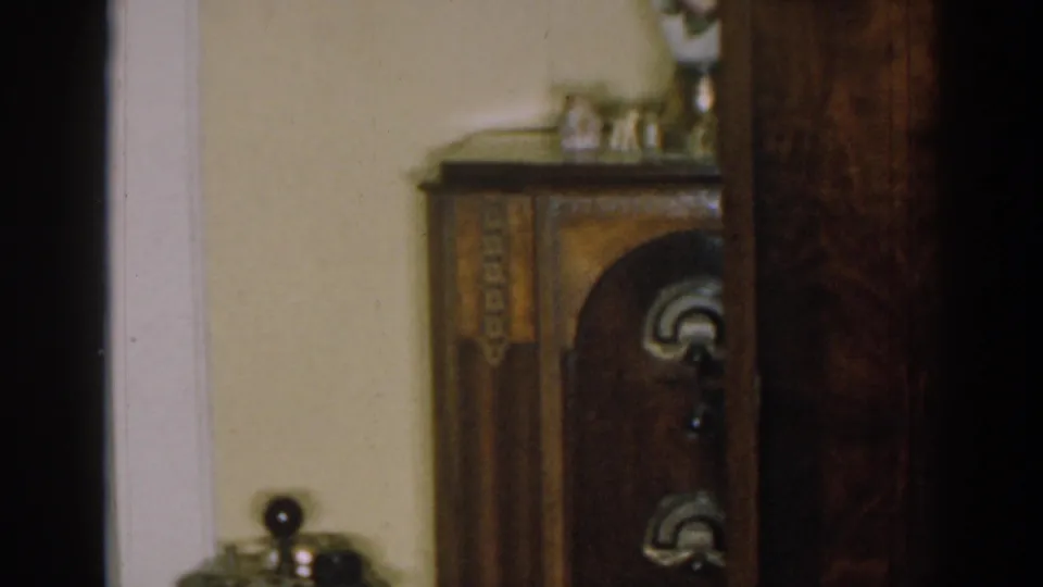 1963 New Jersey home-movie frame showing a low view of the cabinet, ashtray stand, pale wall, and shadowed doorway.