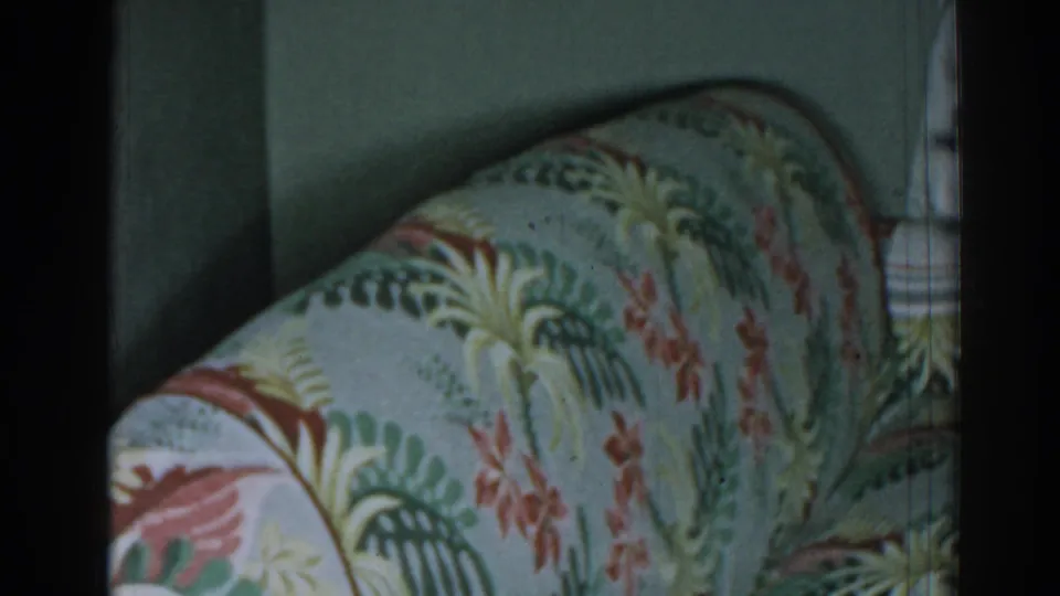 1963 New Jersey home-movie frame showing a close view of a curved tropical-patterned sofa arm near a floral curtain.