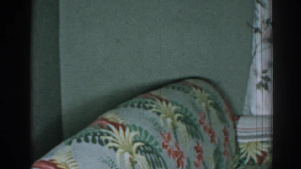 1963 New Jersey interior frame with a patterned sofa back, curved armrest, muted wall, and pale floral curtain.