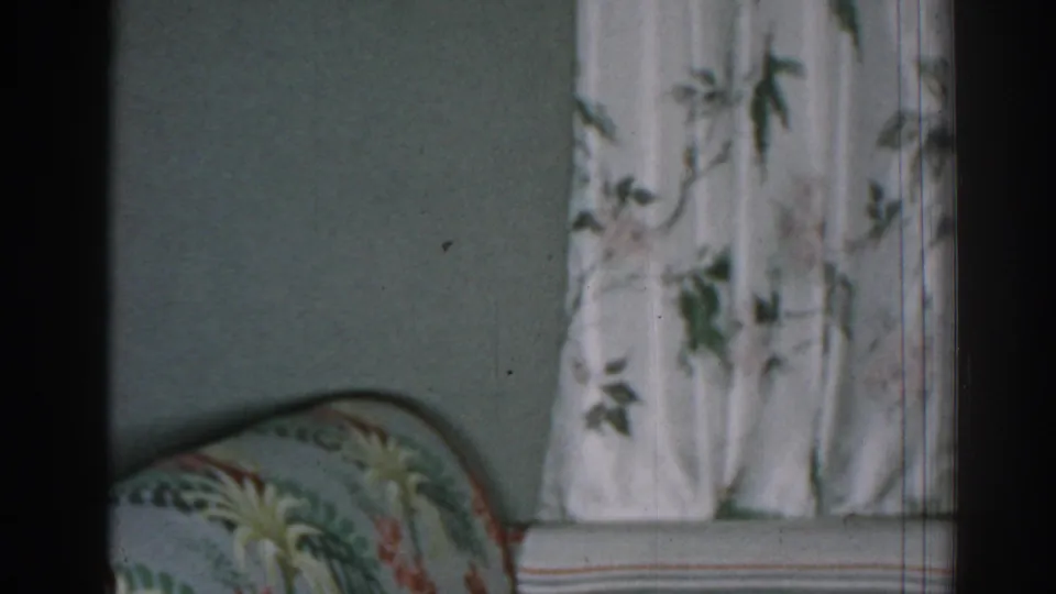 Late-frame 1963 New Jersey living-room view with the patterned sofa arm low in frame and floral curtains beside a muted wall.