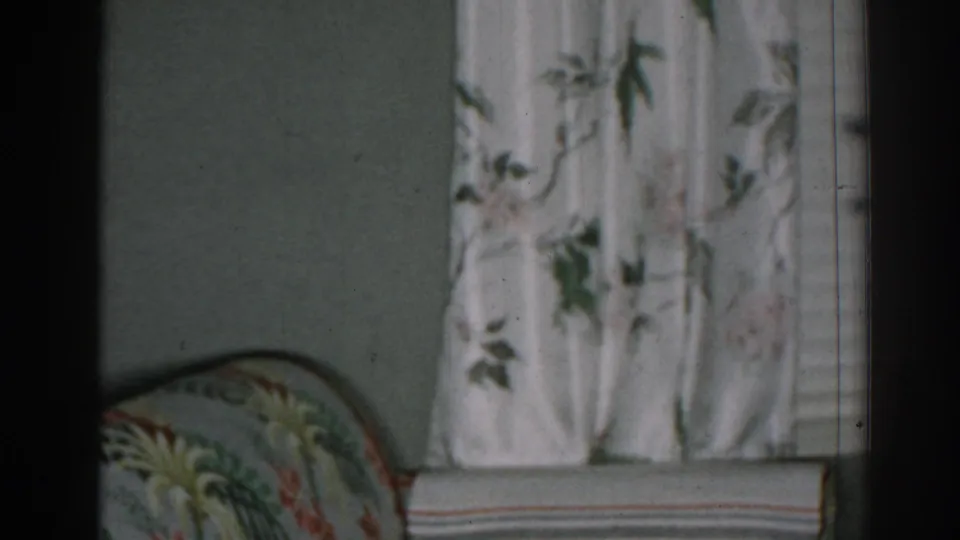 1963 New Jersey home-movie frame showing floral curtains, part of an upholstered armchair, and a pale living-room wall.