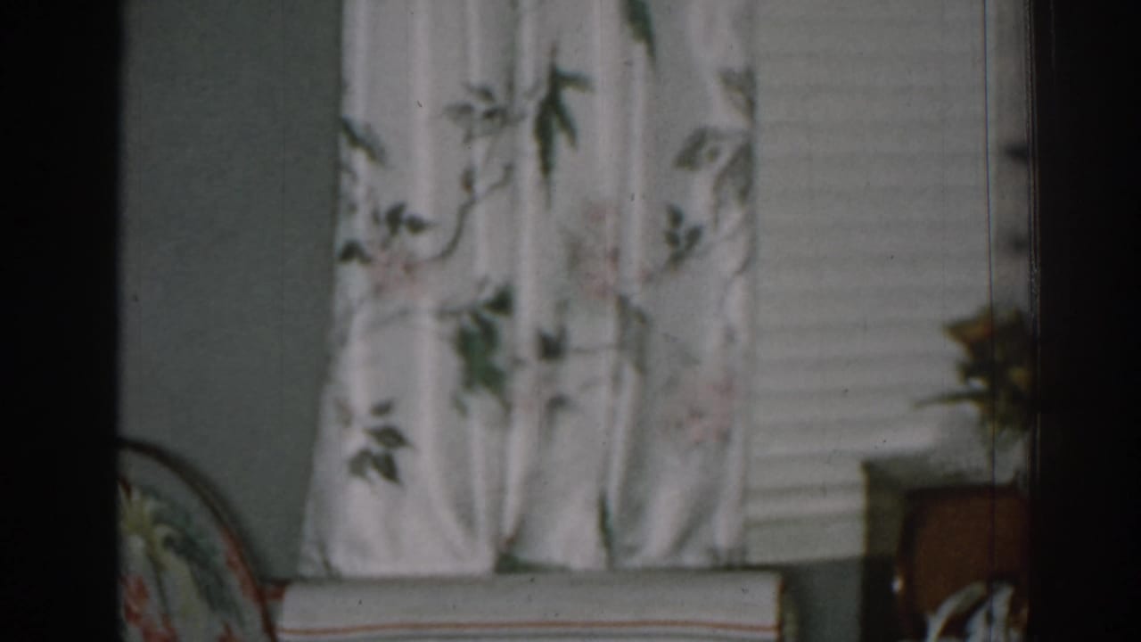 Floral curtain, window blinds, armchair edge, and small table area.