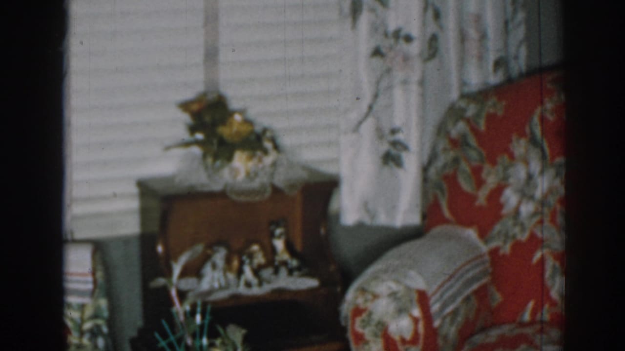 1963 New Jersey living room view showing window blinds, floral curtains, a side table with figurines, and the edge of a red patterned chair.