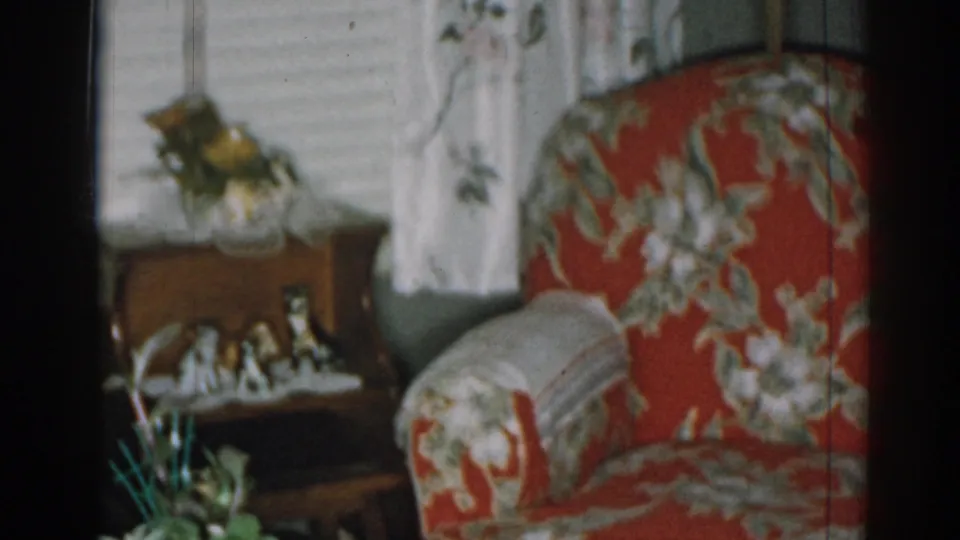 1963 New Jersey home-movie frame with the red floral armchair filling more of the view beside a side table, plants, blinds, and curtains.