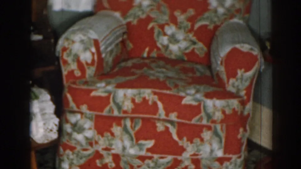 1963 New Jersey living room frame showing a red floral armchair with grey accents, a wooden side table, figurines, flowers, blinds, and curtains.