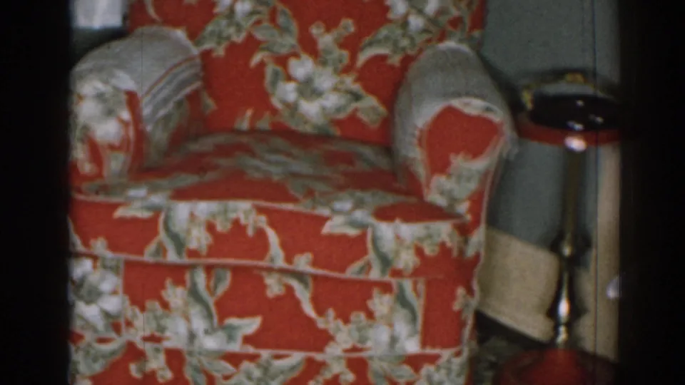 1963 New Jersey home-movie frame showing a red floral armchair next to a small brass-toned side table in a private living room.
