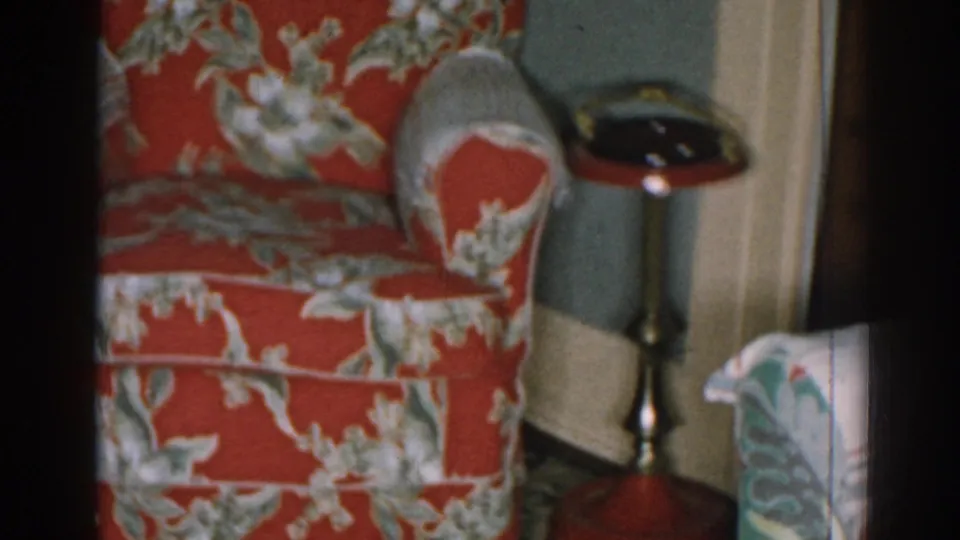 1963 New Jersey interior frame with a red floral armchair, dark-topped pedestal table, pale trim, and greenish wall.