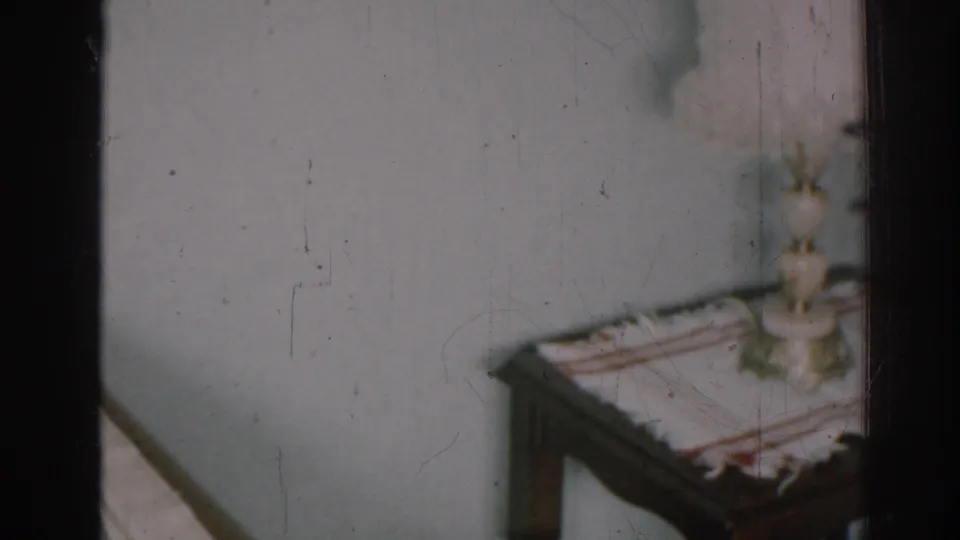 1963 New Jersey home-movie frame showing a nightstand with a striped cloth and lamp beside part of a bed.