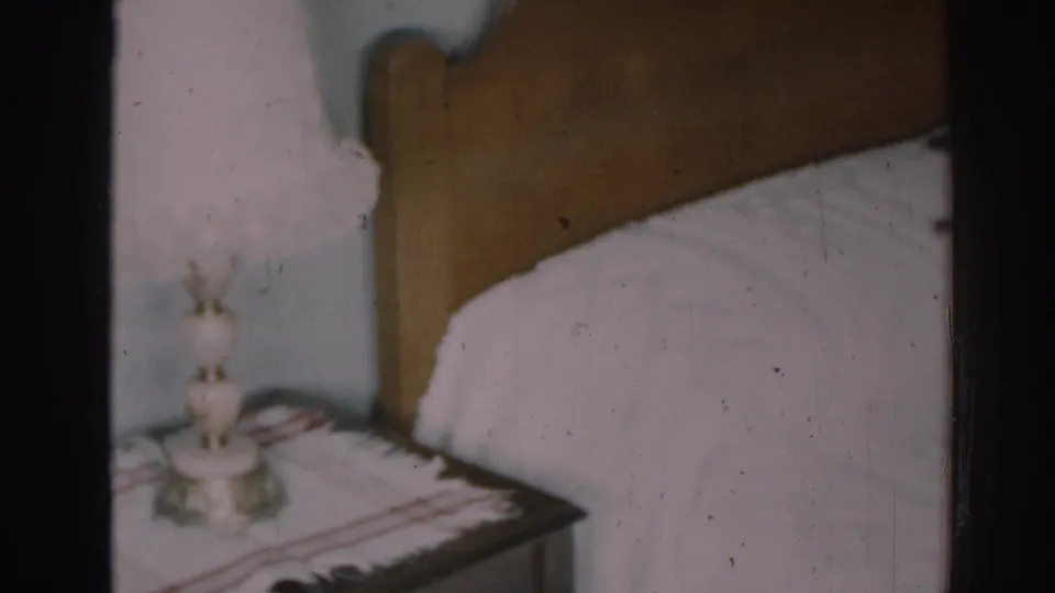 1963 New Jersey home-movie frame showing a wooden headboard, white sheets, and part of the lamp and nightstand.