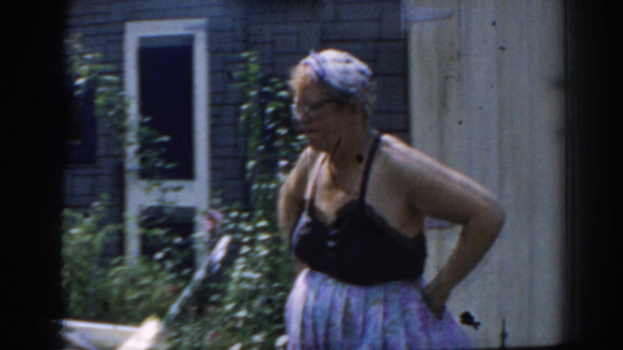 Woman in a dark top, patterned skirt, and headscarf standing near a house exterior.