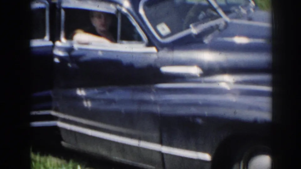 1963 home-movie frame showing the side of a dark blue sedan, chrome trim, open window, and a partially visible occupant.