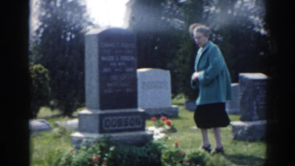 1963 New Jersey 8mm frame showing a woman in a teal jacket and dark skirt standing beside cemetery headstones and flowers.