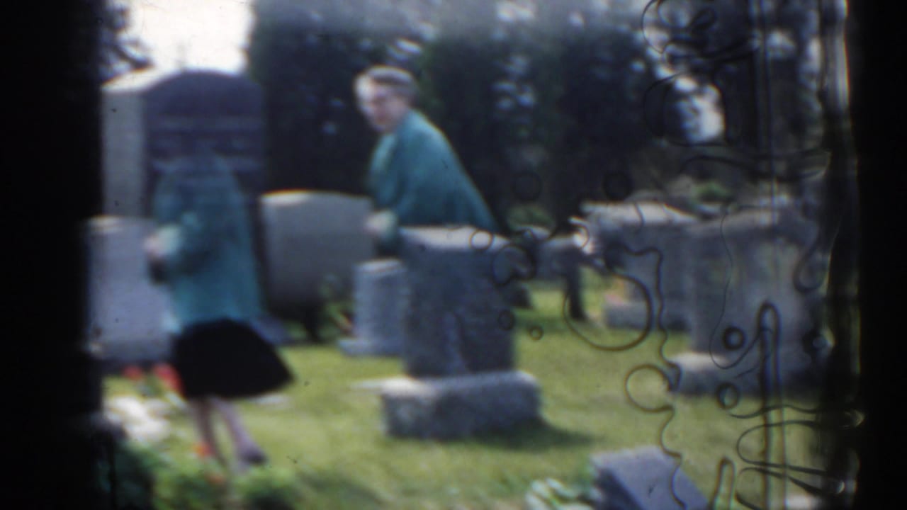 1963 New Jersey home-movie frame of a woman in a teal jacket moving through a cemetery with upright grave markers and grass.