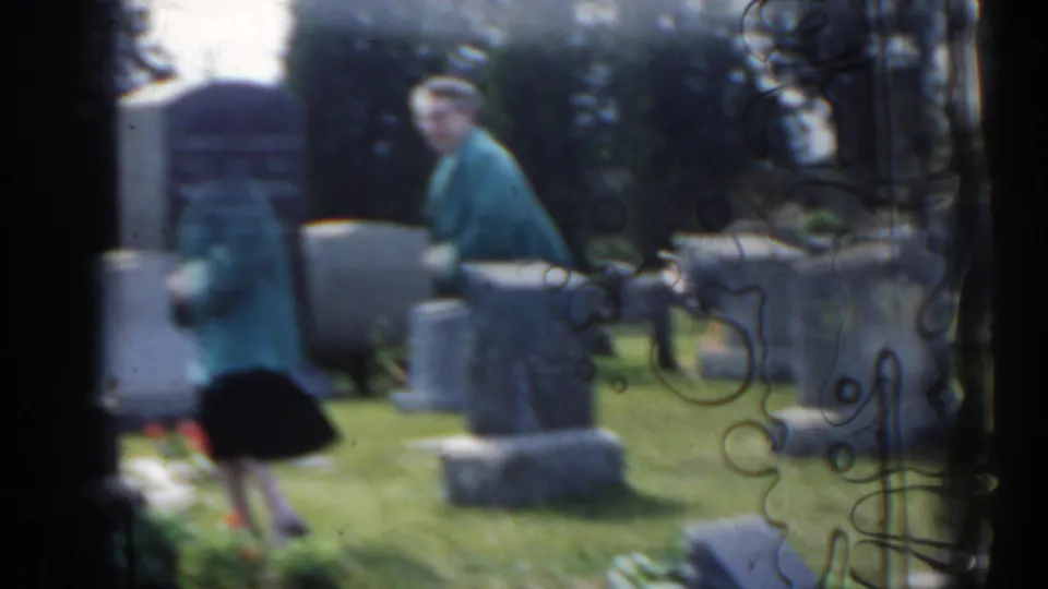 1963 New Jersey home-movie frame of a woman in a teal jacket moving through a cemetery with upright grave markers and grass.