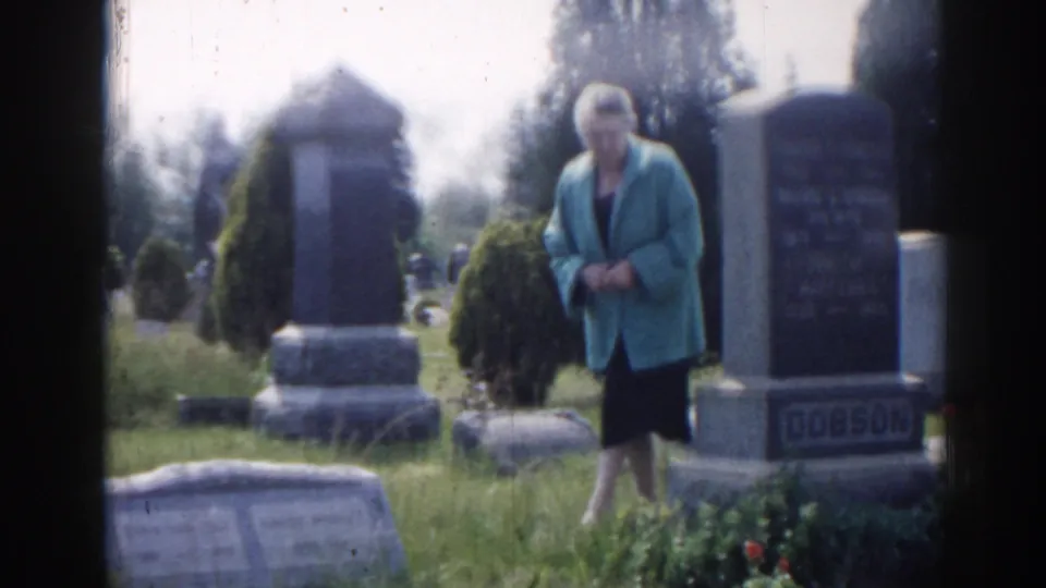 1963 New Jersey frame showing a woman standing near a prominent DOBSON cemetery monument with grass and shrubs around the grave.