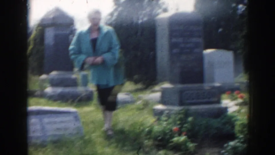 Woman in teal outerwear stands near a large cemetery monument, with red flowers and several headstones visible in the 1963 New Jersey footage.