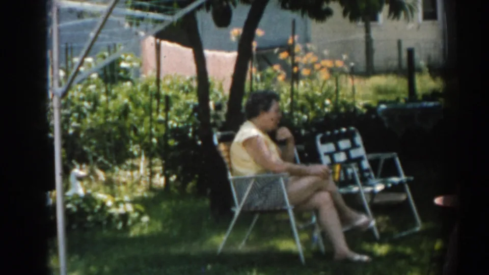 1963 New Jersey 8mm frame showing women in lawn chairs during a backyard conversation, with grass, trees and a house behind them.