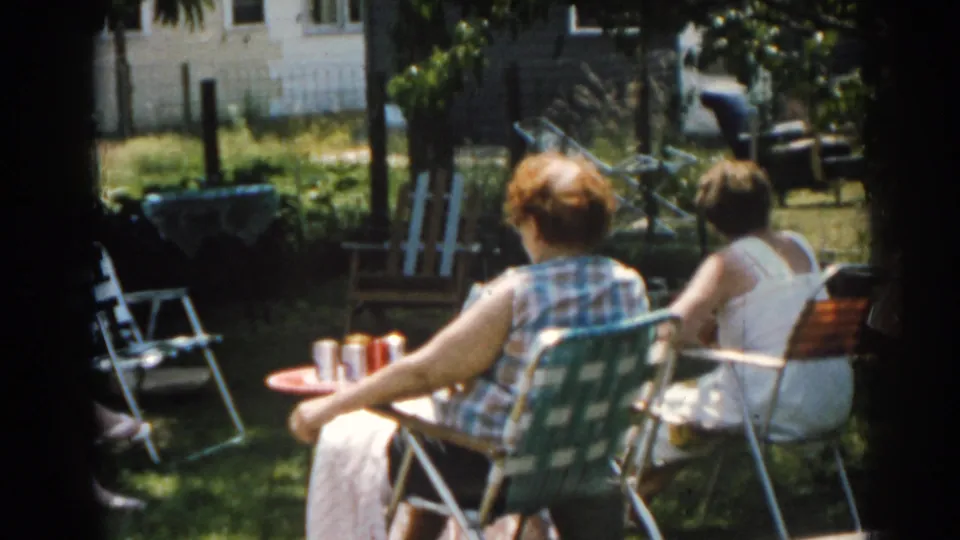 1963 New Jersey backyard frame showing women from behind in folding lawn chairs, a small table with cans and garden activity in the background.