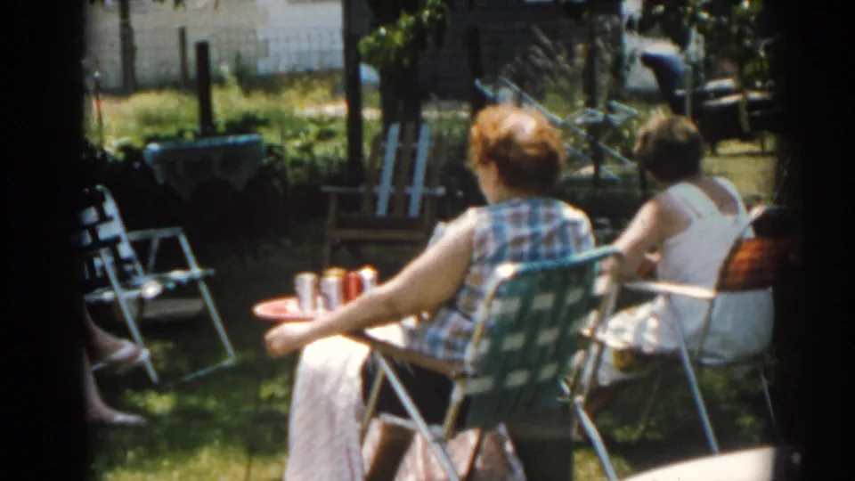 1963 New Jersey home-movie frame of two women seated under trees in a backyard, with a small table, drink cans and garden behind them.