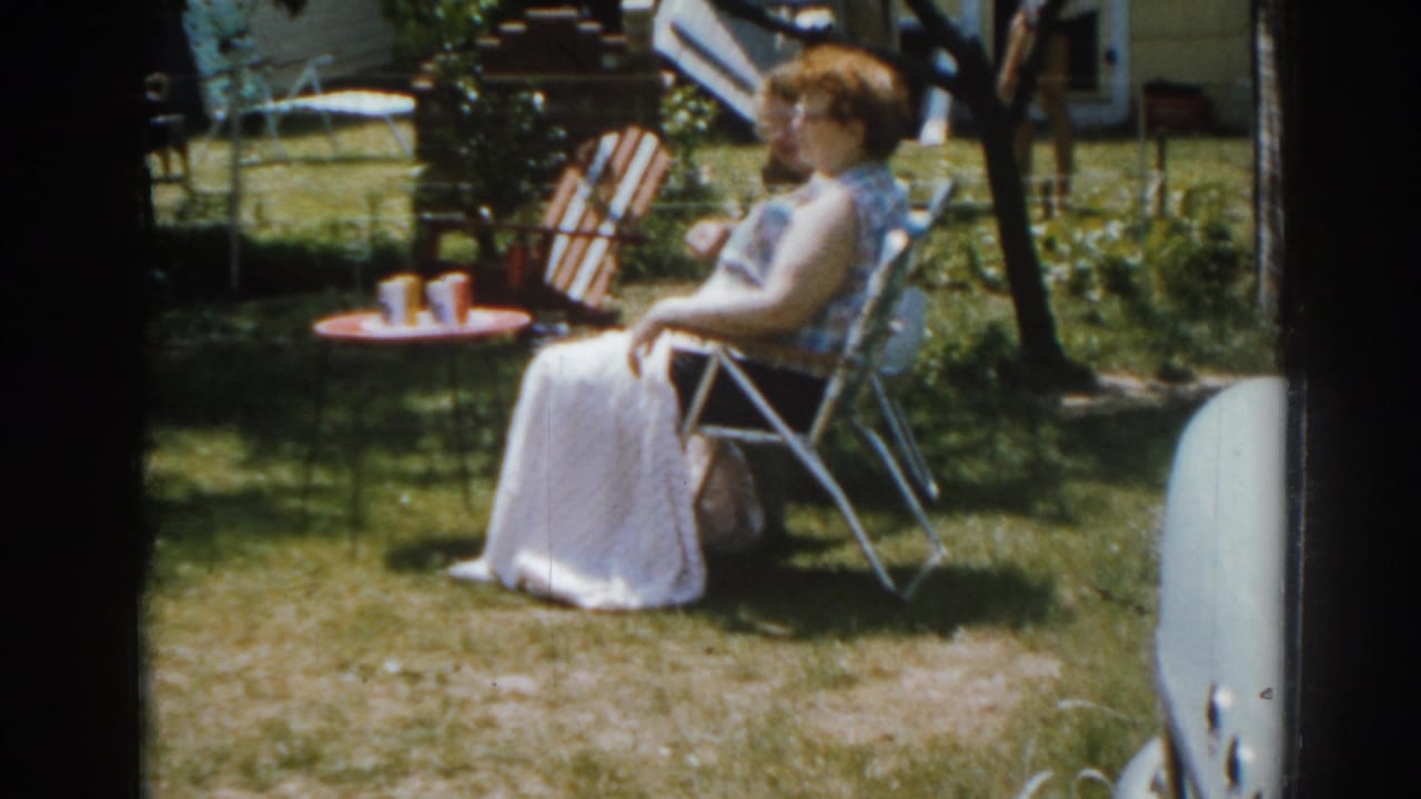 Woman seated in a backyard chair beside a small red table with cups.