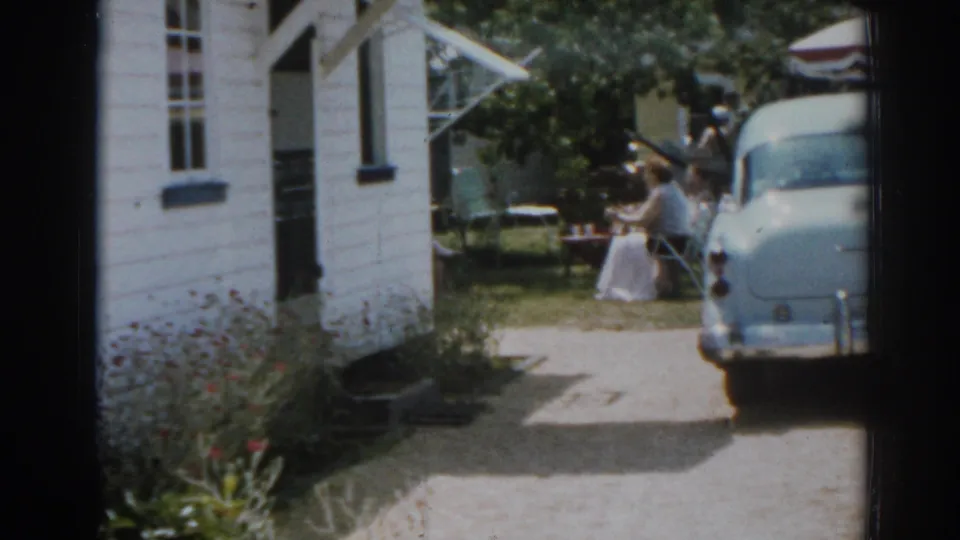 A white house, driveway, rounded vintage car, and seated woman appear together in this 1963 New Jersey backyard home-movie frame.