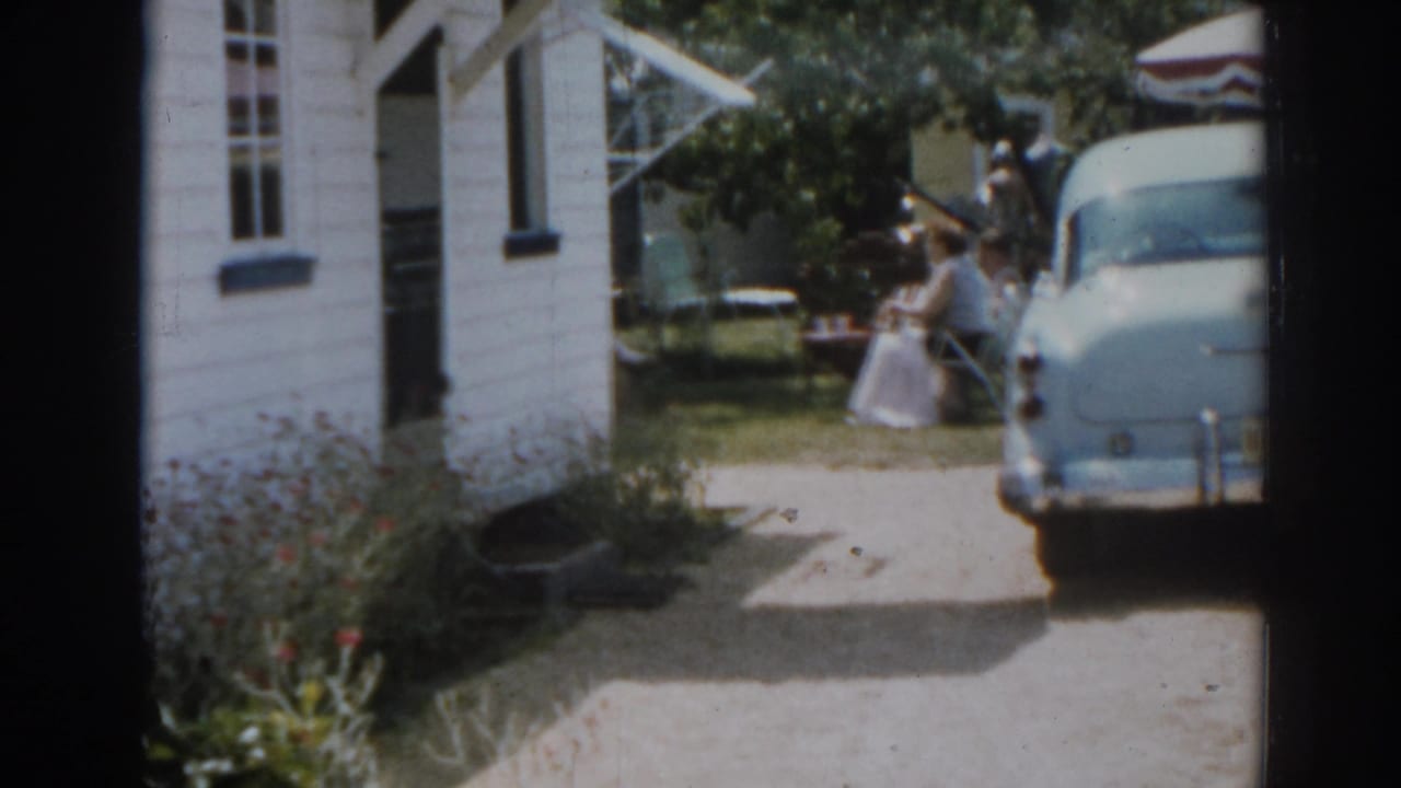 White clapboard house, blue car in driveway, and women near lawn furniture in a sunny yard.