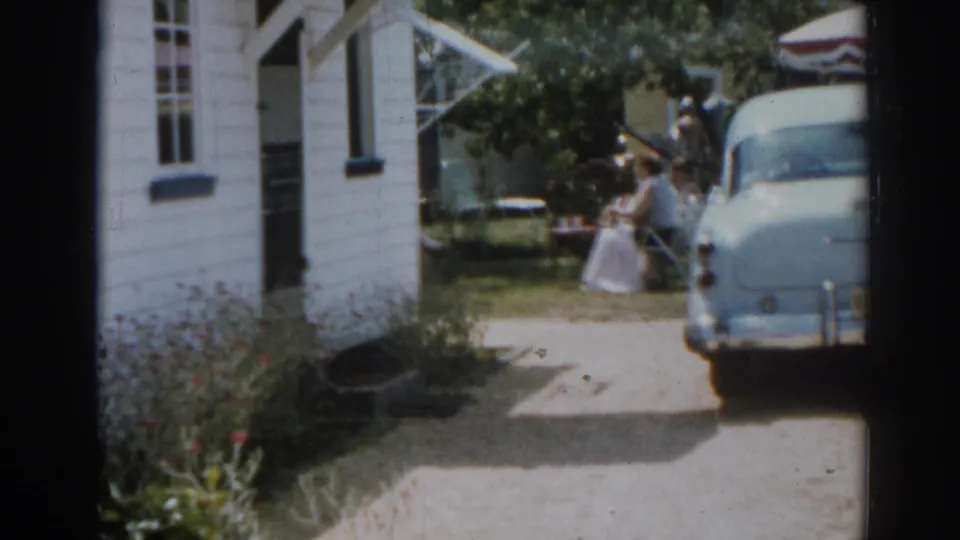 1963 New Jersey home-movie frame showing a white house, blue car, and women gathered near outdoor seating in a sunny backyard.