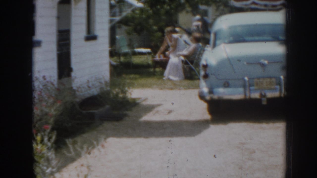 Blue car parked beside a white house as women move near outdoor chairs in the yard.