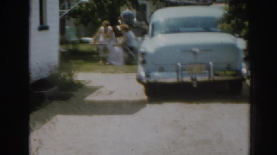 1963 New Jersey home-movie frame showing a blue car in a driveway and women near lawn furniture beside a white house.