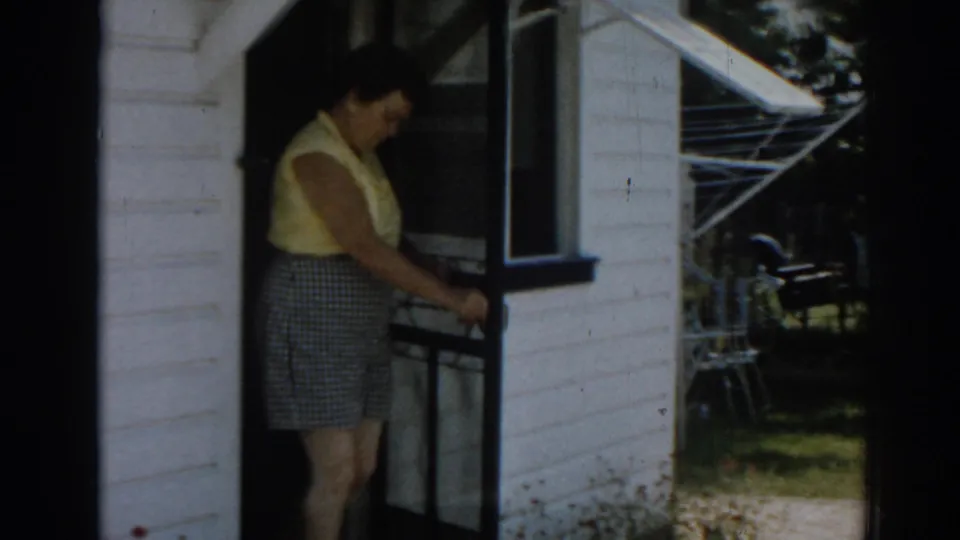 1963 New Jersey home-movie frame showing a woman at the doorway of a white clapboard house with yard furniture and clothesline nearby.