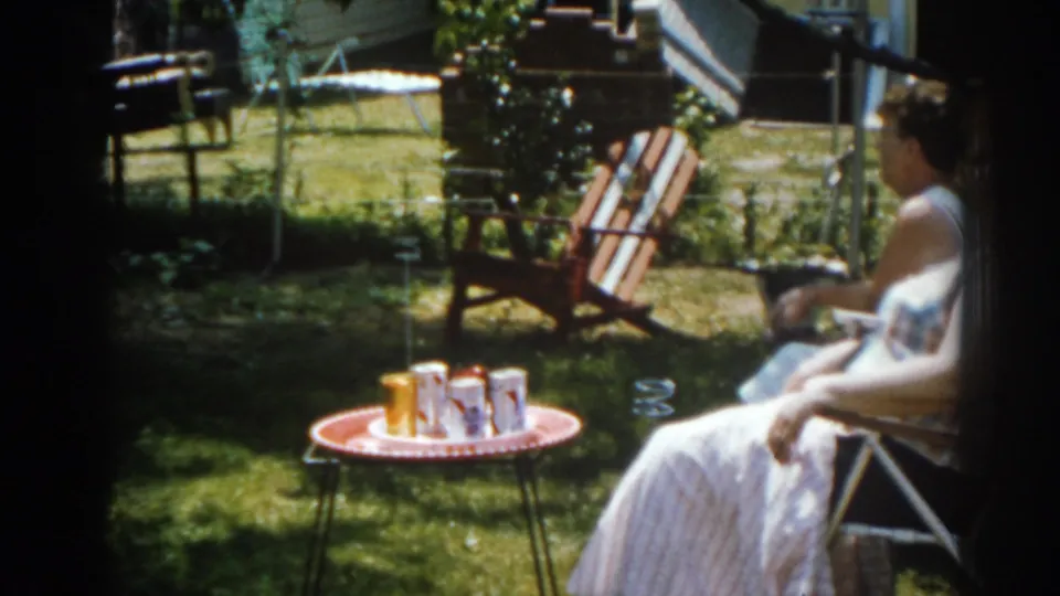 1963 New Jersey backyard frame with a woman seated in a folding chair near drinks on a red patio table.
