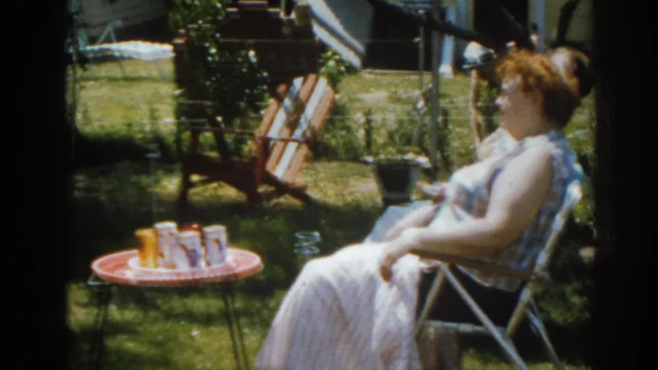 1963 New Jersey home-movie frame showing a woman in profile beside a red table with drinks and backyard seating.