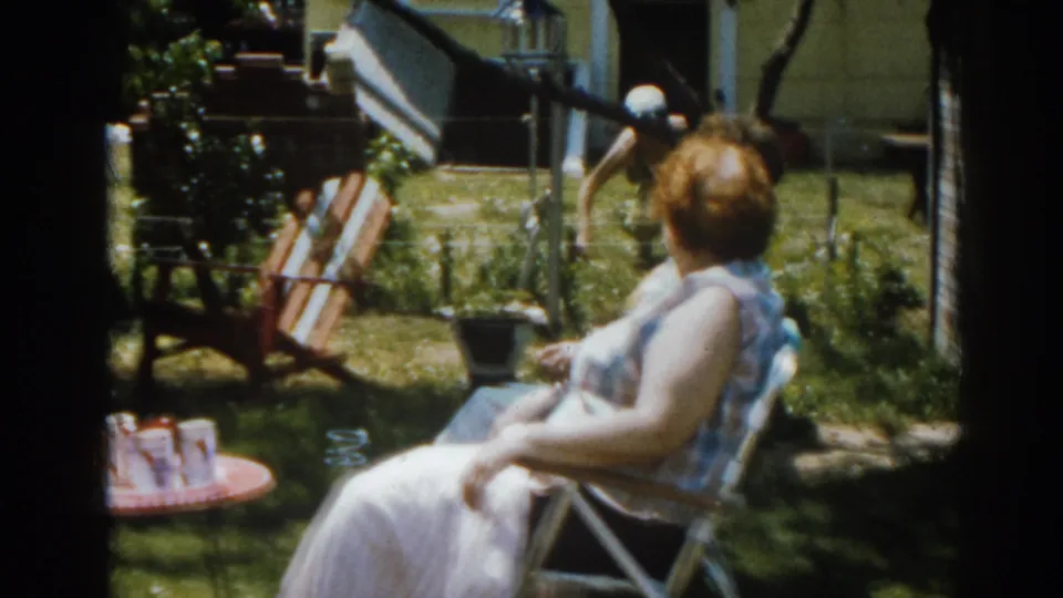 1963 New Jersey 8mm frame with a woman seated in a lawn chair, drinks on a red table, and yard furniture behind her.