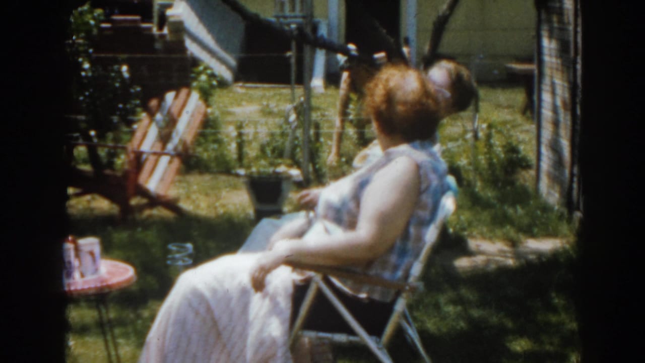 Woman seated in a backyard lawn chair with a wooden chair and small table nearby.