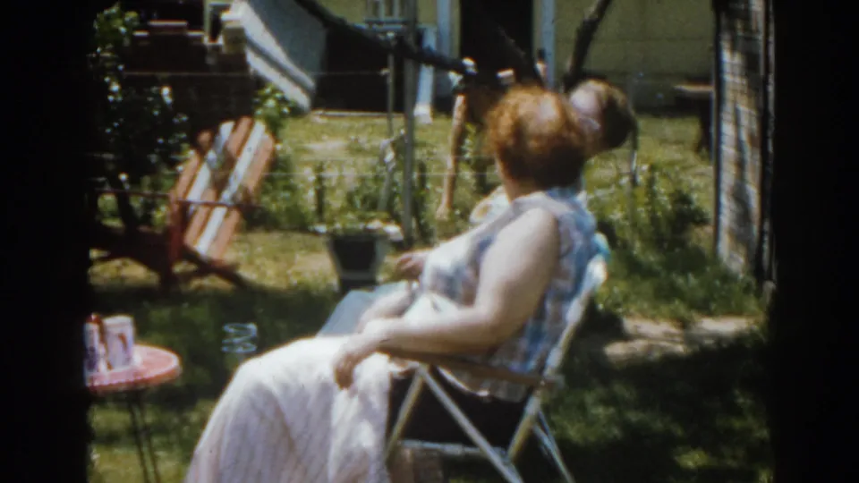1963 New Jersey home-movie frame showing a woman seated in a sunny backyard beside lawn furniture and a house exterior.