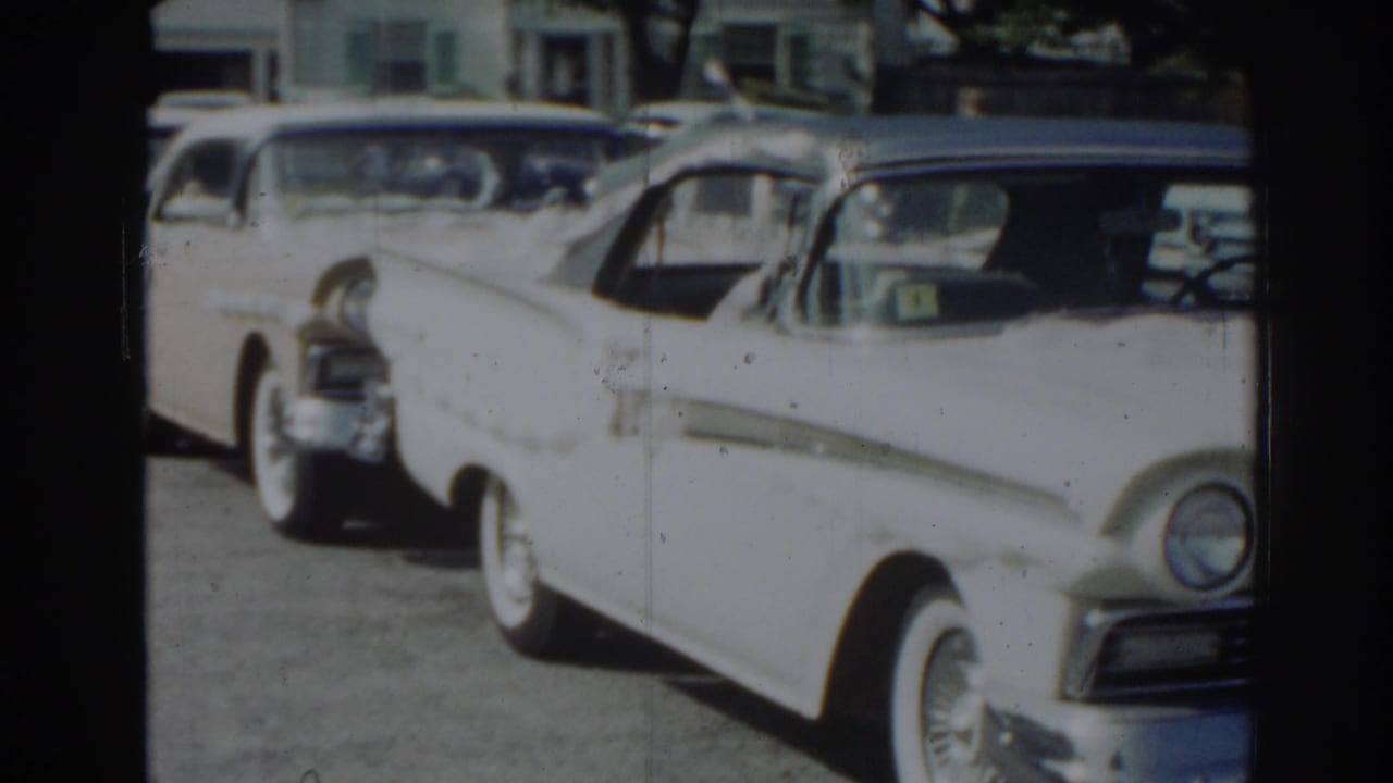 White vintage sedan parked on a suburban street with other cars behind it.