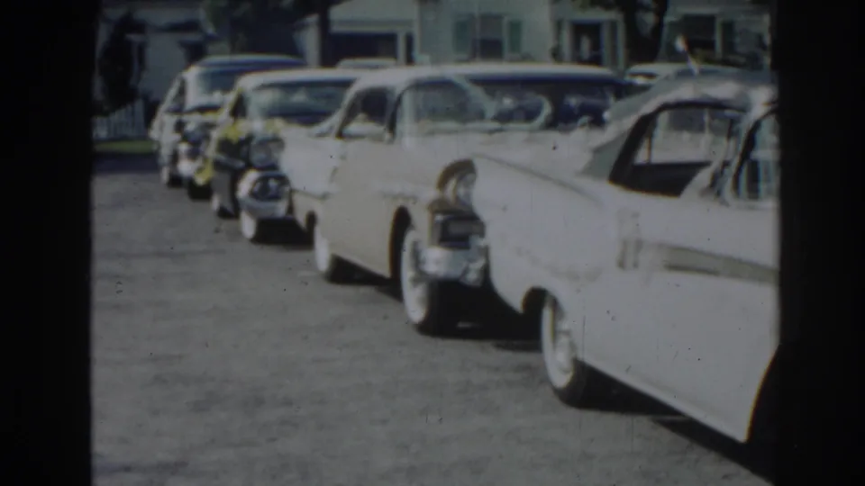 The camera view looks down a quiet New Jersey street lined with several parked 1960s cars and detached homes.