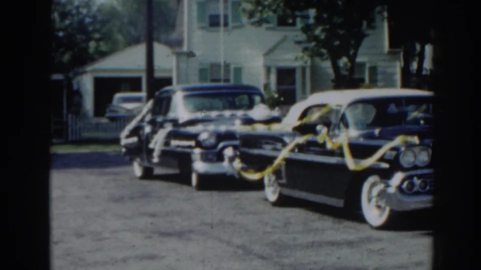 A black ribbon-decorated vintage car stands near other parked cars outside suburban homes in the 1963 New Jersey footage.