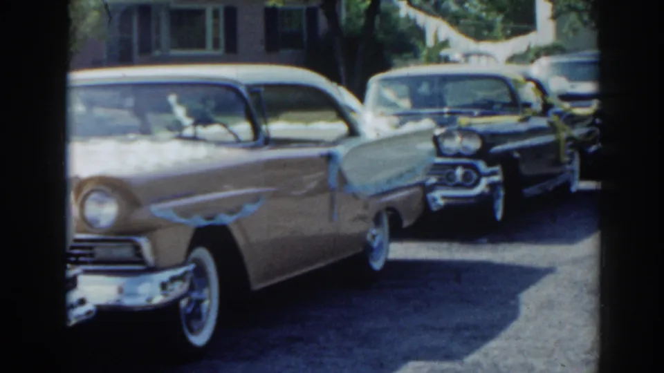 1963 New Jersey home-movie frame showing a light-colored finned sedan beside ribbon-decorated dark classic cars.