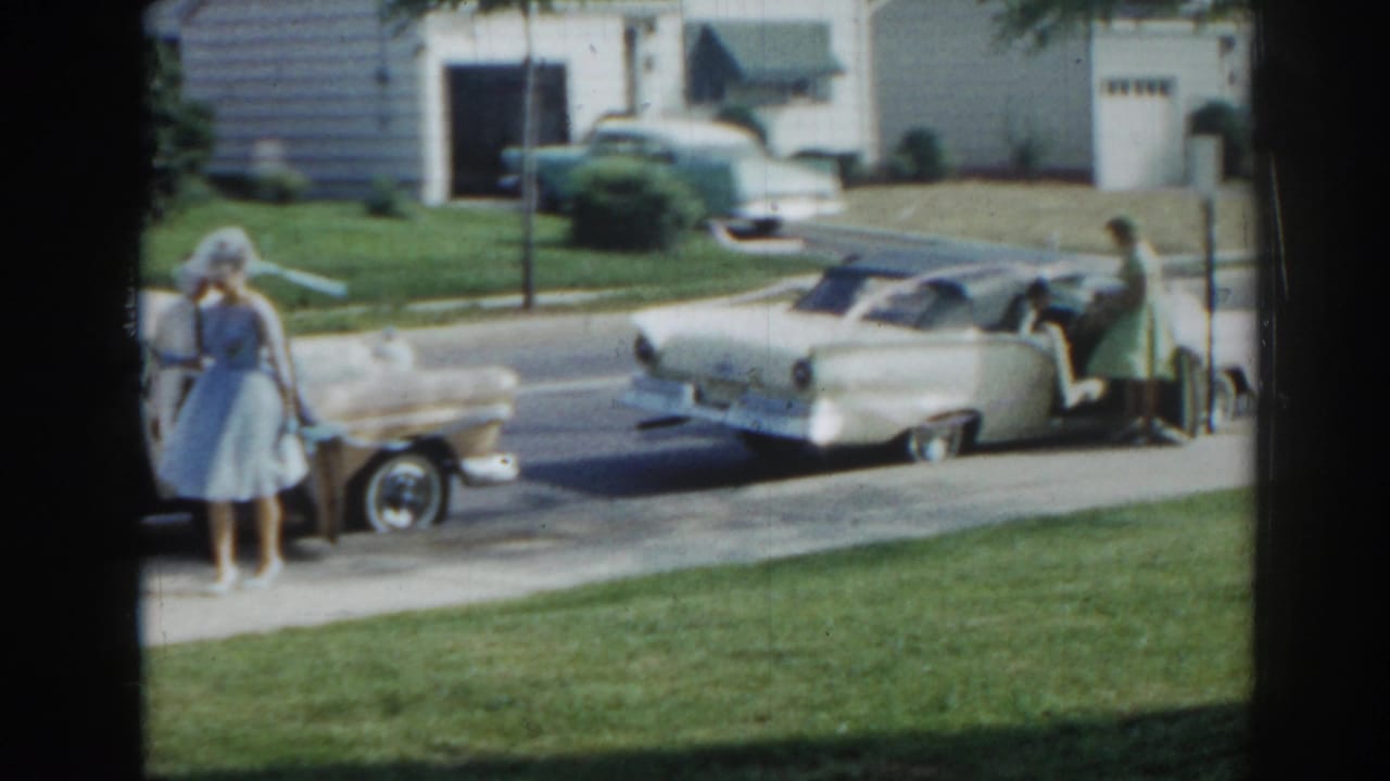 Woman in a blue dress stands near a cream convertible on a suburban street.