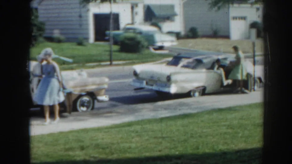 In 1963 New Jersey home-movie footage, a woman in a blue dress and hat stands by parked mid-century cars on a residential street.