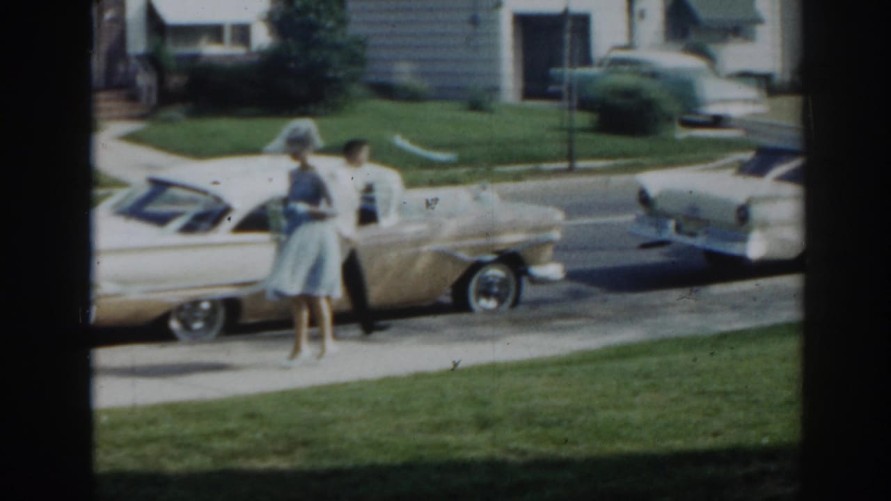 Woman in blue stands beside a cream convertible as a man stands near the passenger side.