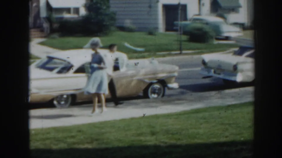 A woman in a blue dress and hat stands beside a cream convertible, with a man nearby on a suburban New Jersey street in 1963.