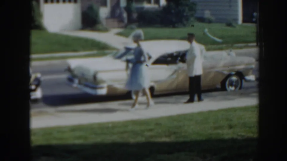 A woman in a light blue dress walks along the sidewalk past a cream convertible as a man stands beside it in 1963 New Jersey.