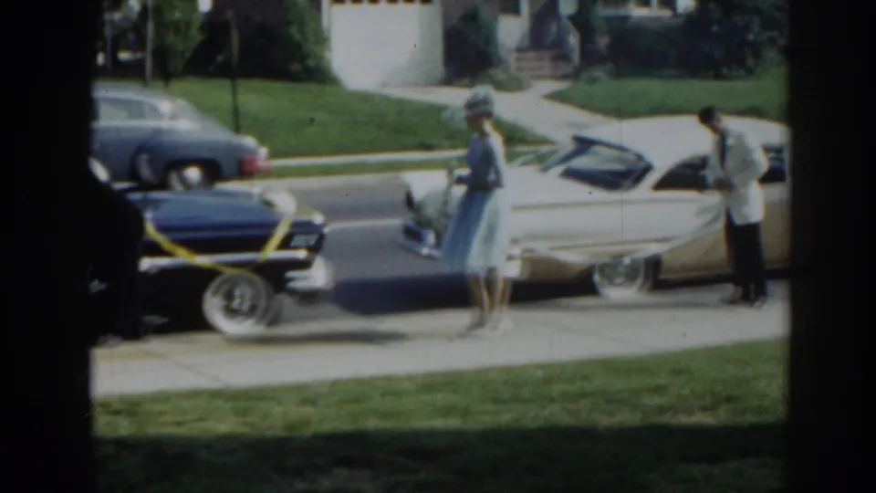 A woman in a blue dress and hat walks between parked vintage cars as a man stands by the cream convertible in New Jersey, 1963.