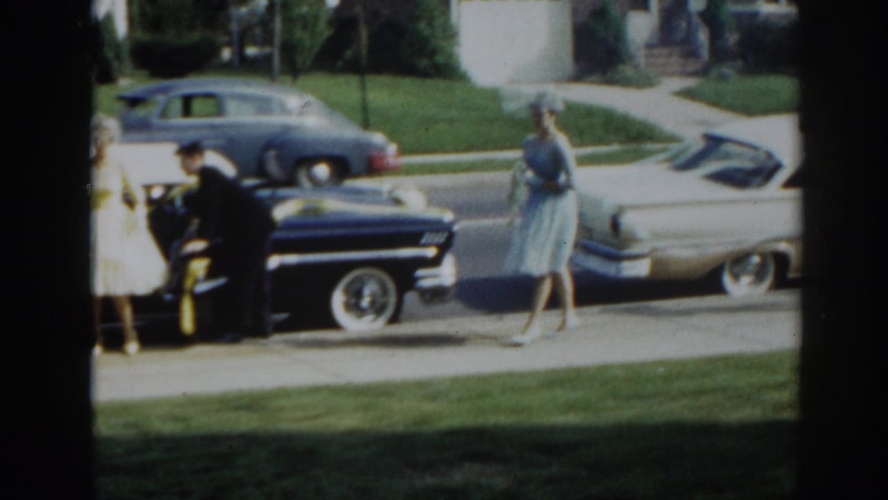 1963 New Jersey home-movie frame showing a man near a ribboned black sedan while women in dresses stand on the sidewalk.