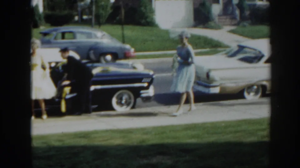 1963 New Jersey home-movie frame showing a man near a ribboned black sedan while women in dresses stand on the sidewalk.